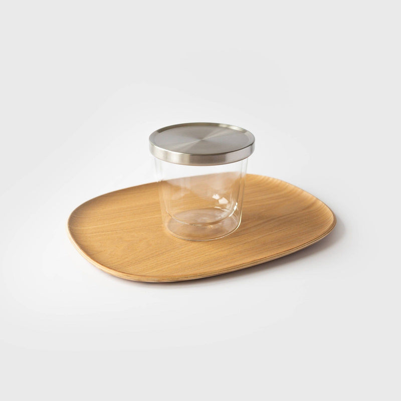 A Kinto USA double-wall glass cup with a metal lid sits elegantly on a round wooden tray against a plain backdrop, providing both style and heat-resistant functionality.
