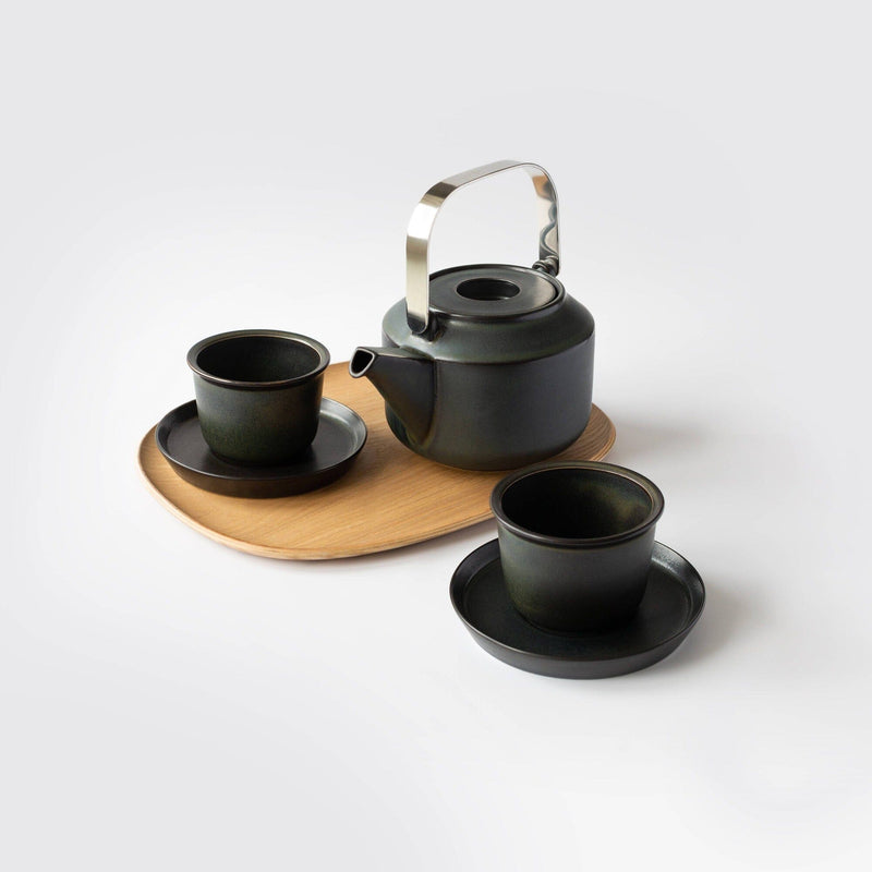 A refined black teapot with a metallic handle sits on a light wood tray, accompanied by two elegant black teacups and saucers. This sophisticated arrangement echoes the Rishi Tea & Botanicals set's timeless charm and minimalist design.